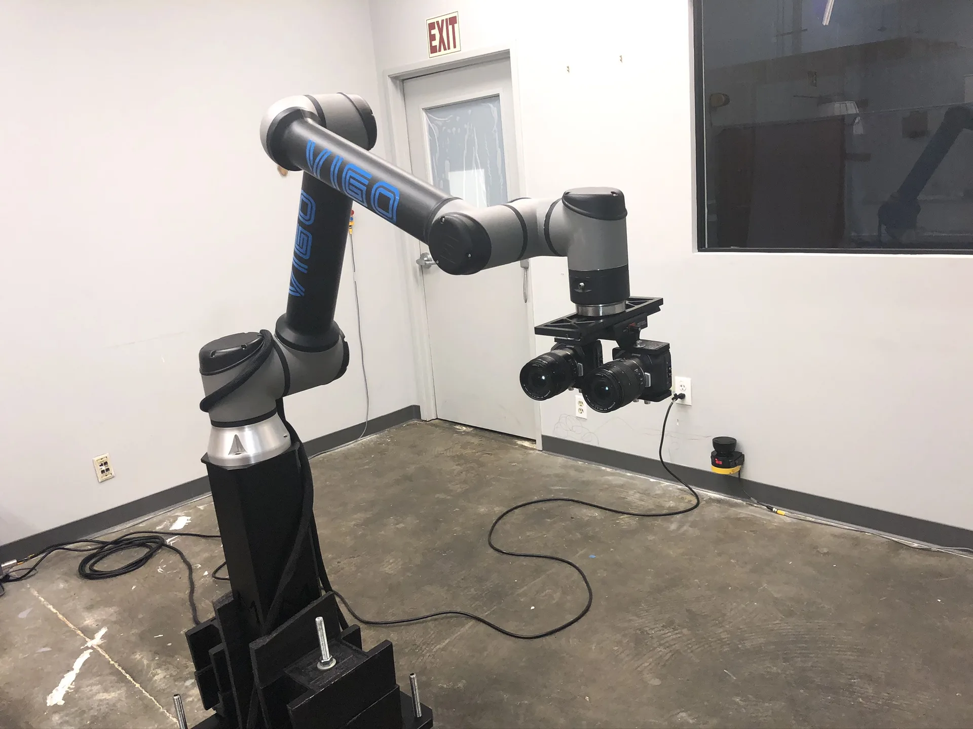 A robotic arm mounted on a black pedestal reaching across a room, with the stereo camera rig at its end.