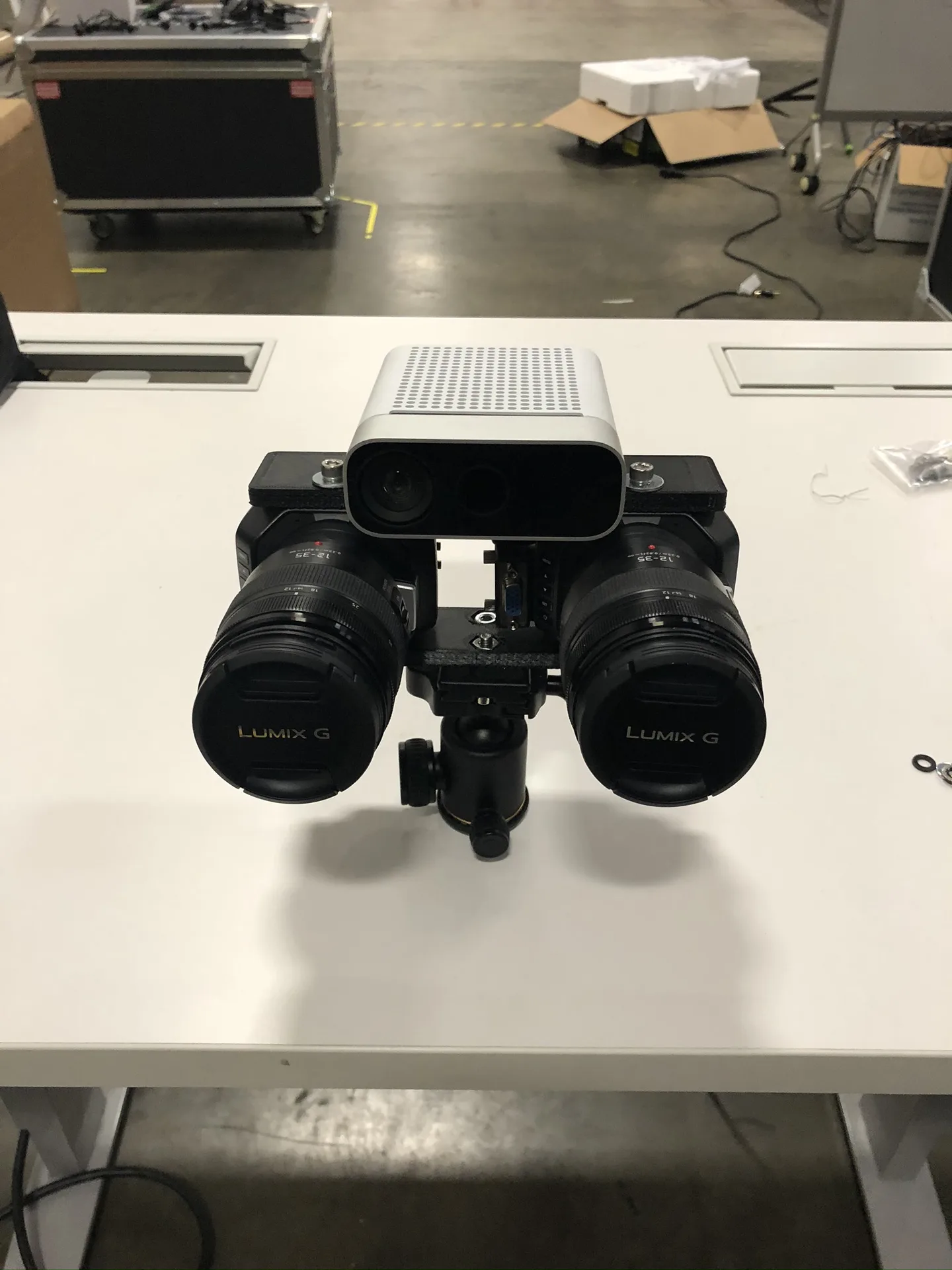 The stereo pair on a ball head on a desk in a warehouse, with a grey RGB and depth sensor mounted above the lenses.